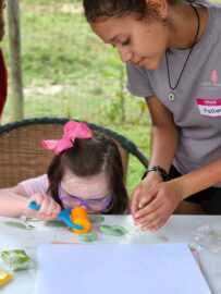 Young girl sitting at art table while doing a craft with a staff person assisting her.