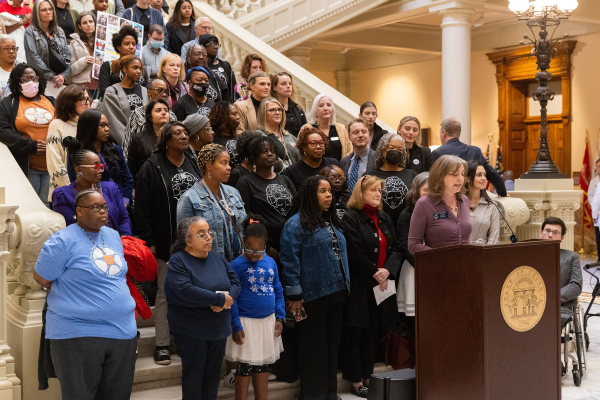 A woman is giving a speech at a seminar, and people are standing on the stairs behind her, listening.